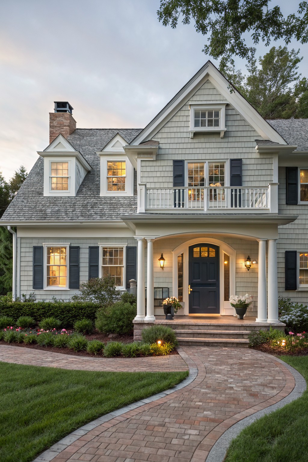 Front view of a two-story light gray shingled house with white trim, gabled roof, columned porch, dark wood entry door, brick pathway, landscaped yard, and warm interior lights visible through windows at dusk.