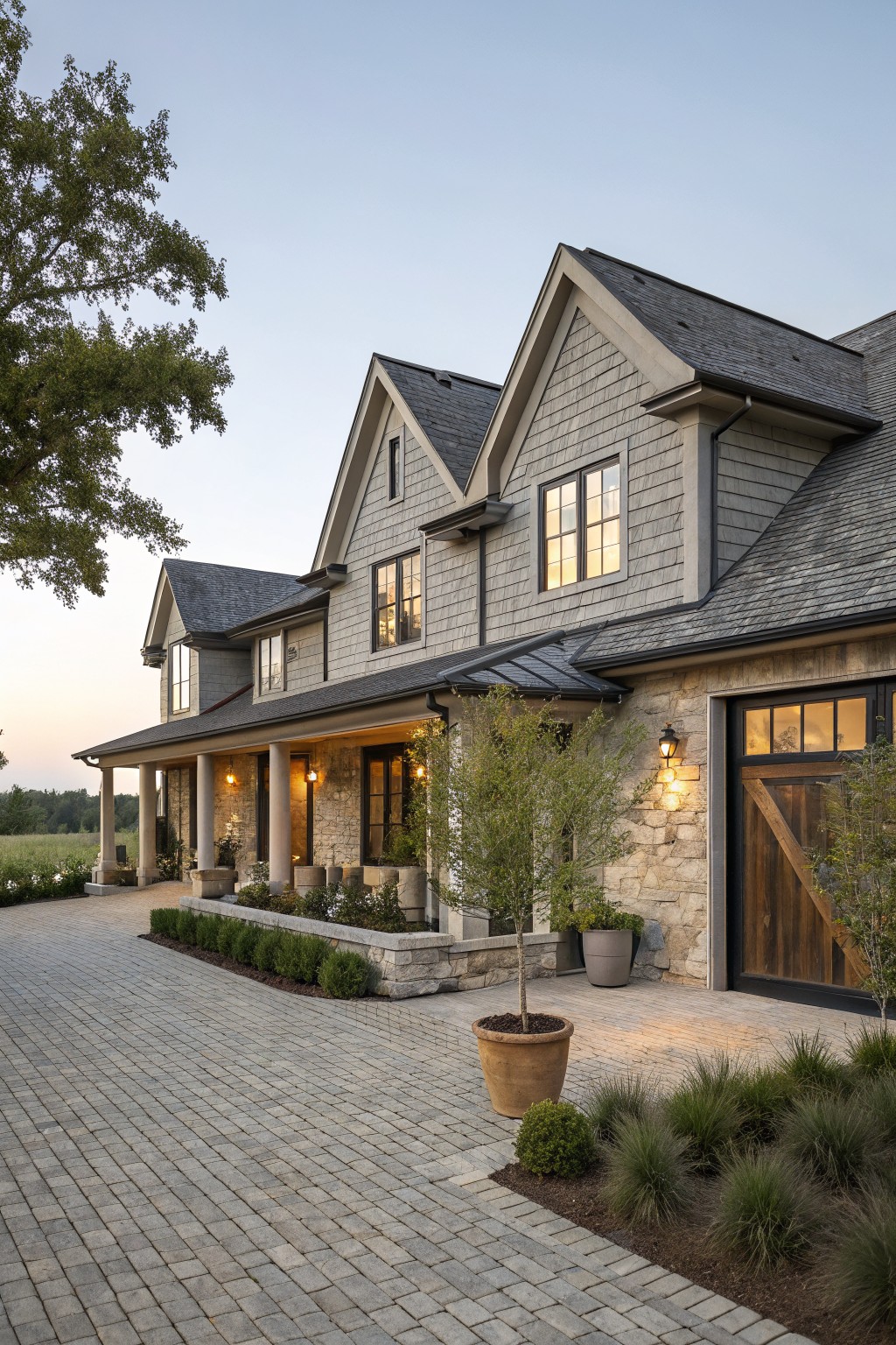 Two-story house exterior featuring light gray shingle siding, gabled roofs, wraparound porch with stone pillars, wood garage door, paver driveway, and low plantings at dusk.
