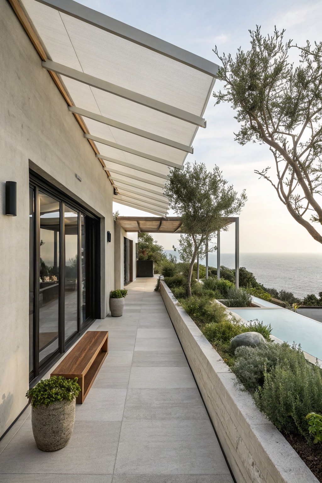 Exterior view of a light gray stucco house wall adjacent to a tiled walkway under a white fabric-covered metal canopy, featuring a wooden bench, potted plant, raised beds with shrubs and boulders, olive trees, and an infinity-edge pool overlooking the ocean.