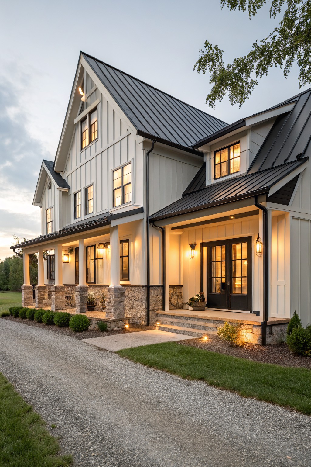 Two-story modern farmhouse with light board-and-batten siding, black metal roof, wrap-around porch supported by stone columns, glowing windows, boxwood shrubs along the foundation, gravel driveway, and lawn at evening.