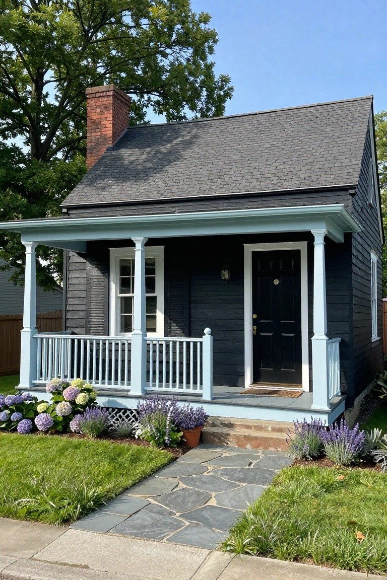 A small black shingled Cape Cod house with a turquoise porch supported by white columns, black front door, white-framed windows, red brick chimney, and front yard landscaping of hydrangeas, lavender plants, and a stone walkway.