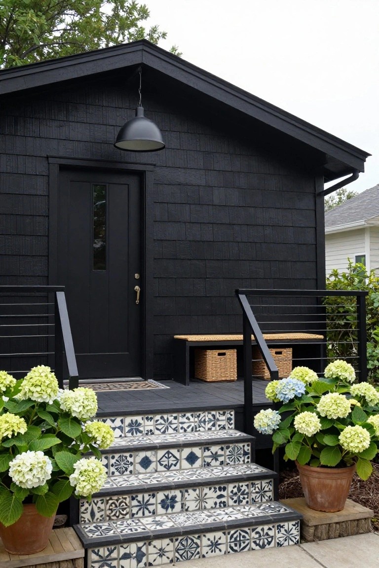 Black shingled shed exterior featuring a black front door with slim window, black metal railings, wooden benches with woven baskets, blue-and-white patterned tile steps, and large potted hydrangeas in terracotta pots.