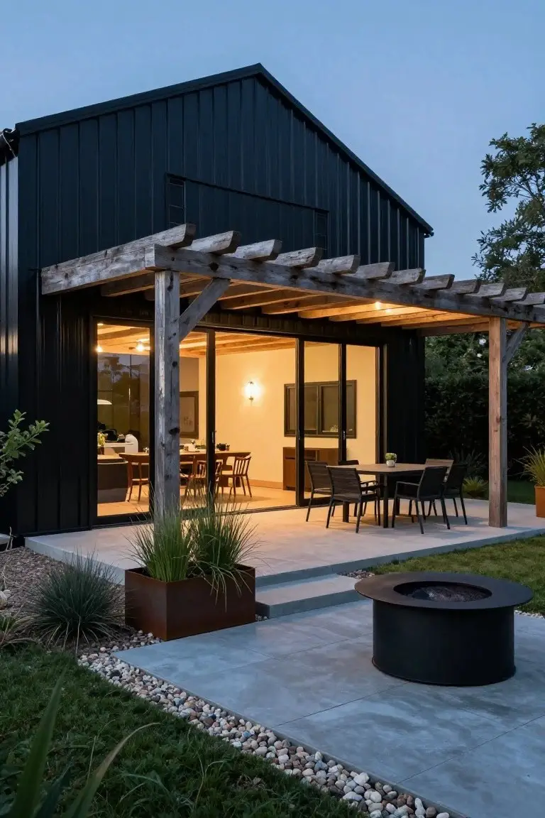 Black metal-clad barn-style house with wooden pergola-covered patio, sliding glass doors to indoor dining area, outdoor table and chairs, potted plants, fire pit, and gravel-edged lawn at dusk.