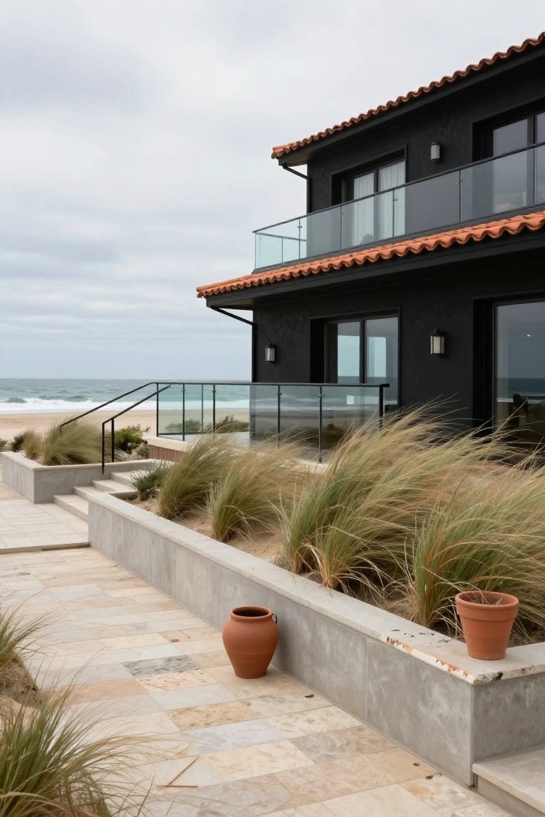 Side exterior view of a two-story black stucco house with terracotta tile roof, glass balcony railings, concrete retaining walls planted with dune grass, terracotta pots, and stairs leading toward a beach with ocean waves in the background.