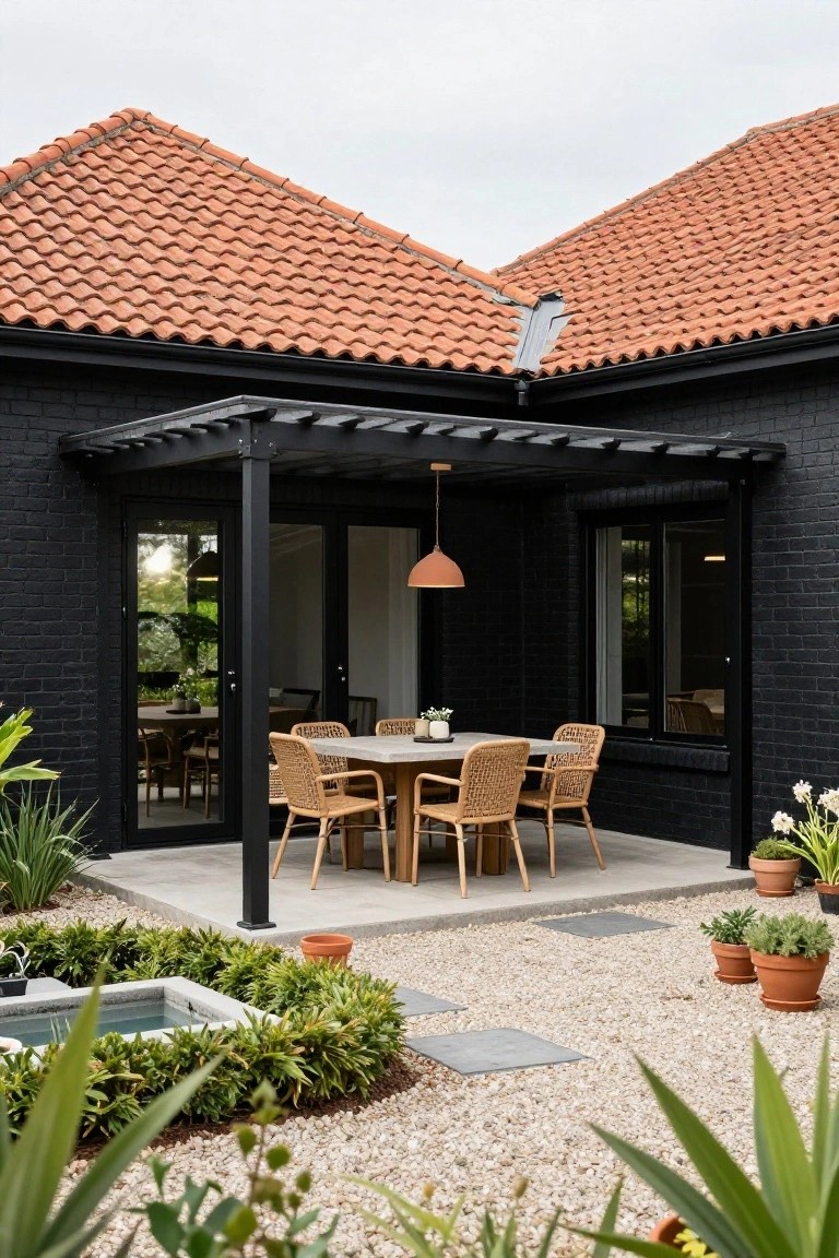Corner exterior of black brick house with terracotta tile roofs, black steel pergola sheltering round table with woven chairs on gravel patio amid plants and small fountain.