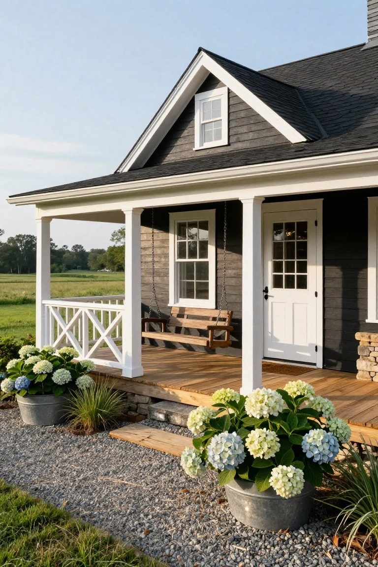 Front view of a dark gray shingle house with gabled roof, white-trimmed porch featuring columns, X-rail balustrade, hanging wooden swing, white paneled door, and potted white hydrangeas on gravel beside wooden steps.