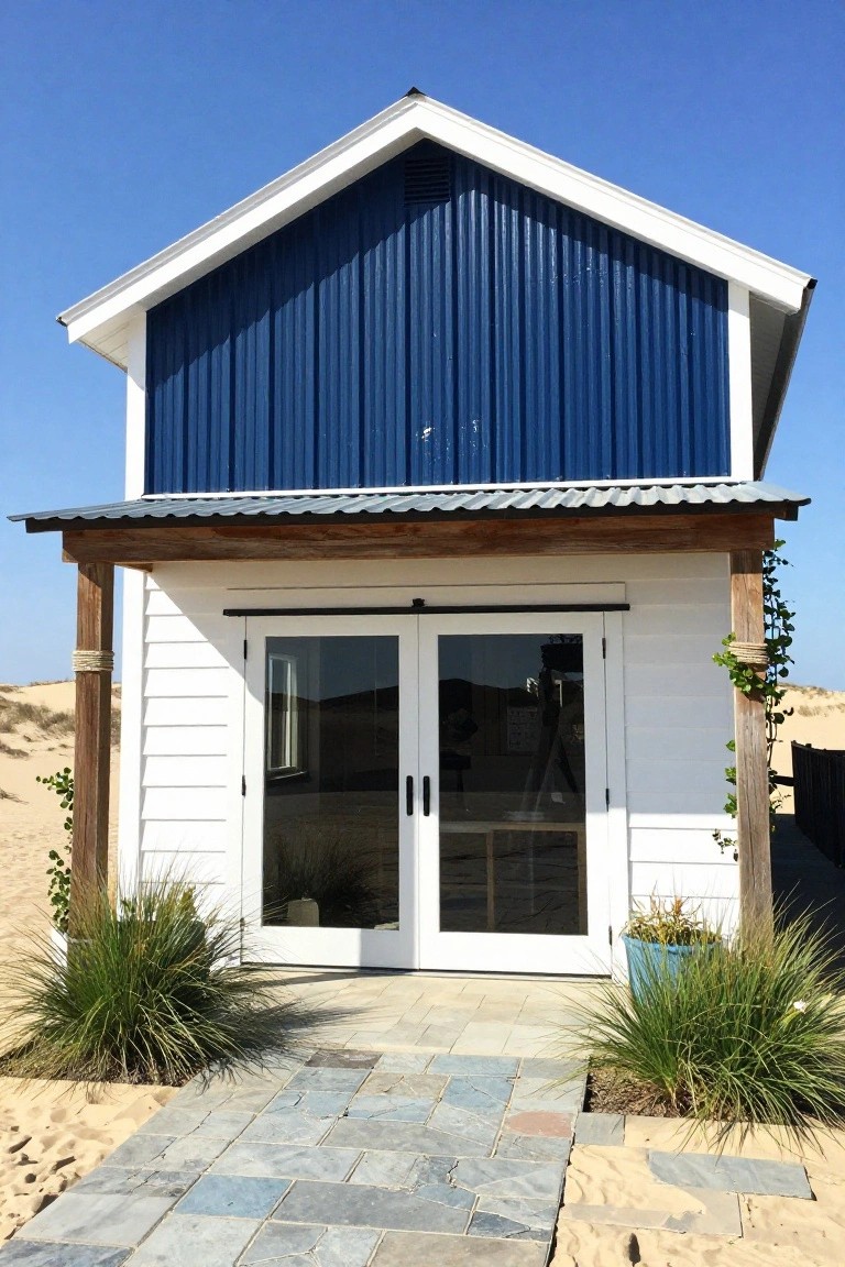A small beach house with blue corrugated metal siding on the upper section, white siding and large glass doors on the lower entry level, wooden posts with rope accents, and potted grasses amid sand dunes.
