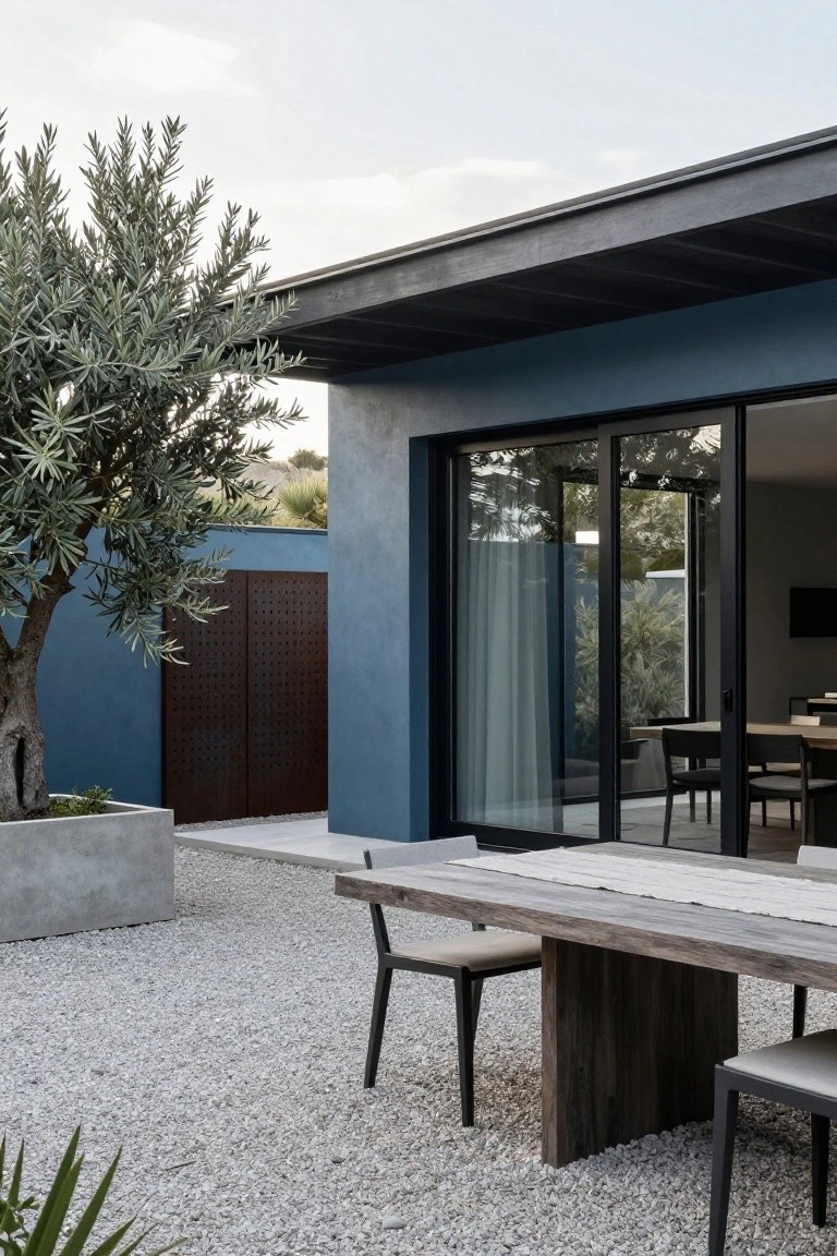 Blue rendered house wall with dark overhanging roof and sliding glass doors open to an interior dining area, adjacent gravel courtyard with wooden dining table and chairs, olive tree in concrete planter, and agave plants.