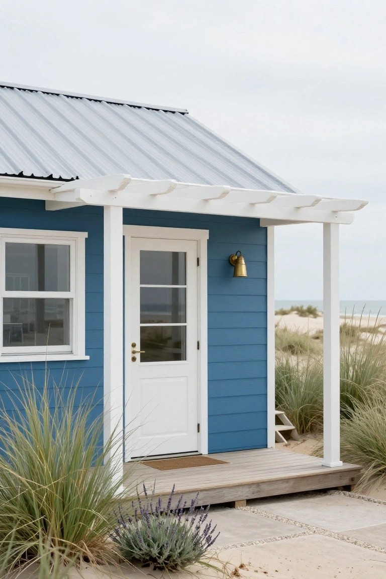Blue clapboard beach house with white trim, front door, pergola, porch steps, and lantern, surrounded by sand dunes, sea grass, and lavender plants near the ocean.