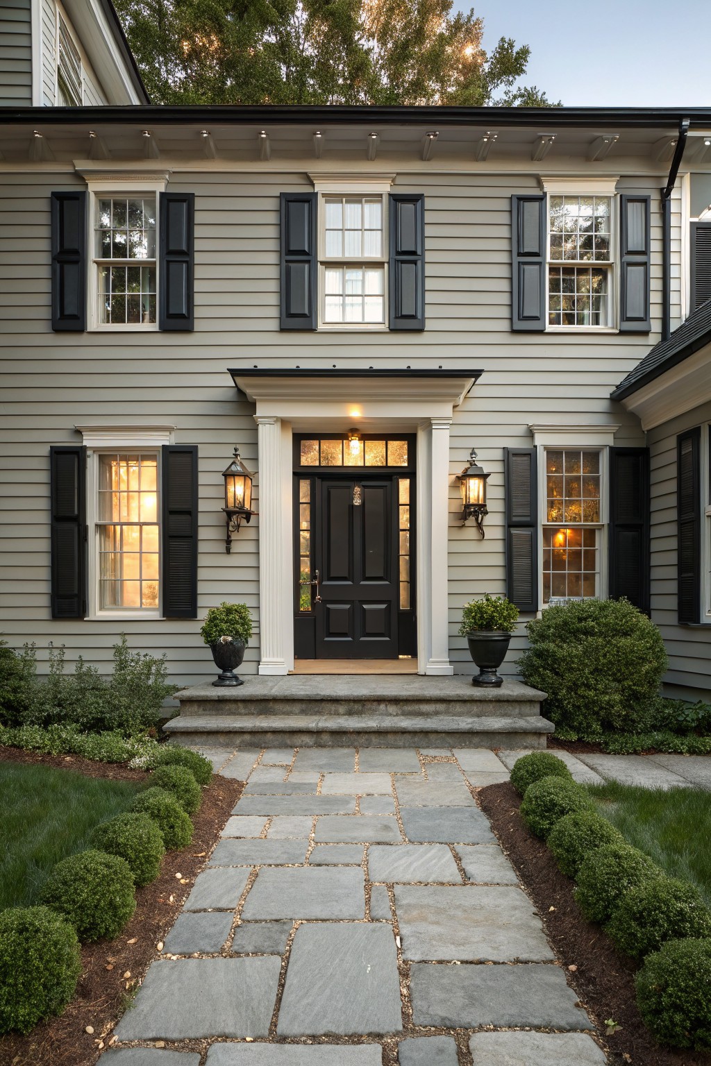 Two-story light gray clapboard house with black shutters on multi-pane windows, black front door under white pediment and columns with black lanterns, stone steps and pathway bordered by boxwood shrubs and grass.