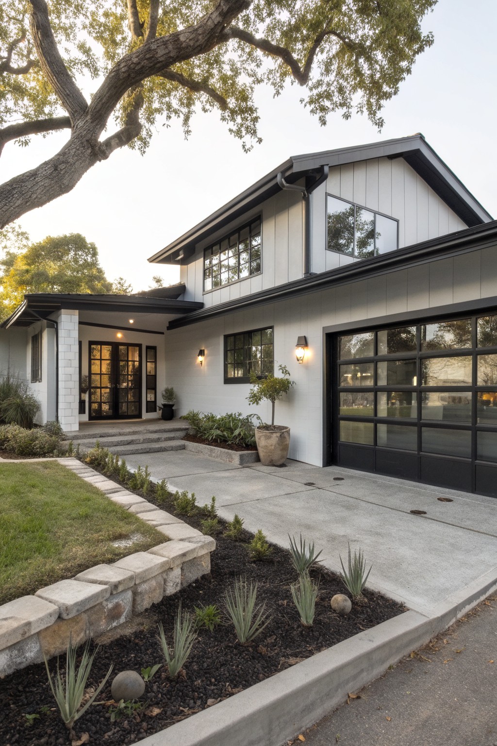 A two-story modern house with light gray vertical siding, black trim on large windows and doors, black-framed glass garage door, covered entry porch, stone retaining wall with agave plants, and oak trees nearby.