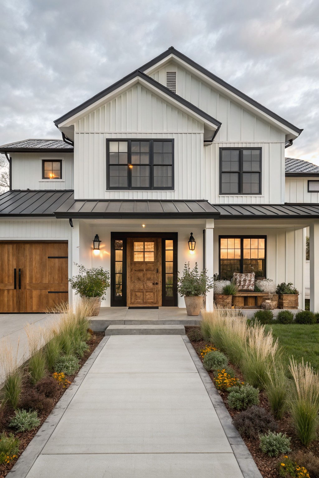 Front view of a two-story house with white vertical board-and-batten siding, black standing-seam metal roof over the main structure and porch, black-framed windows, wooden front door and garage door, lanterns on the porch, and a concrete walkway edged with grasses and plants.