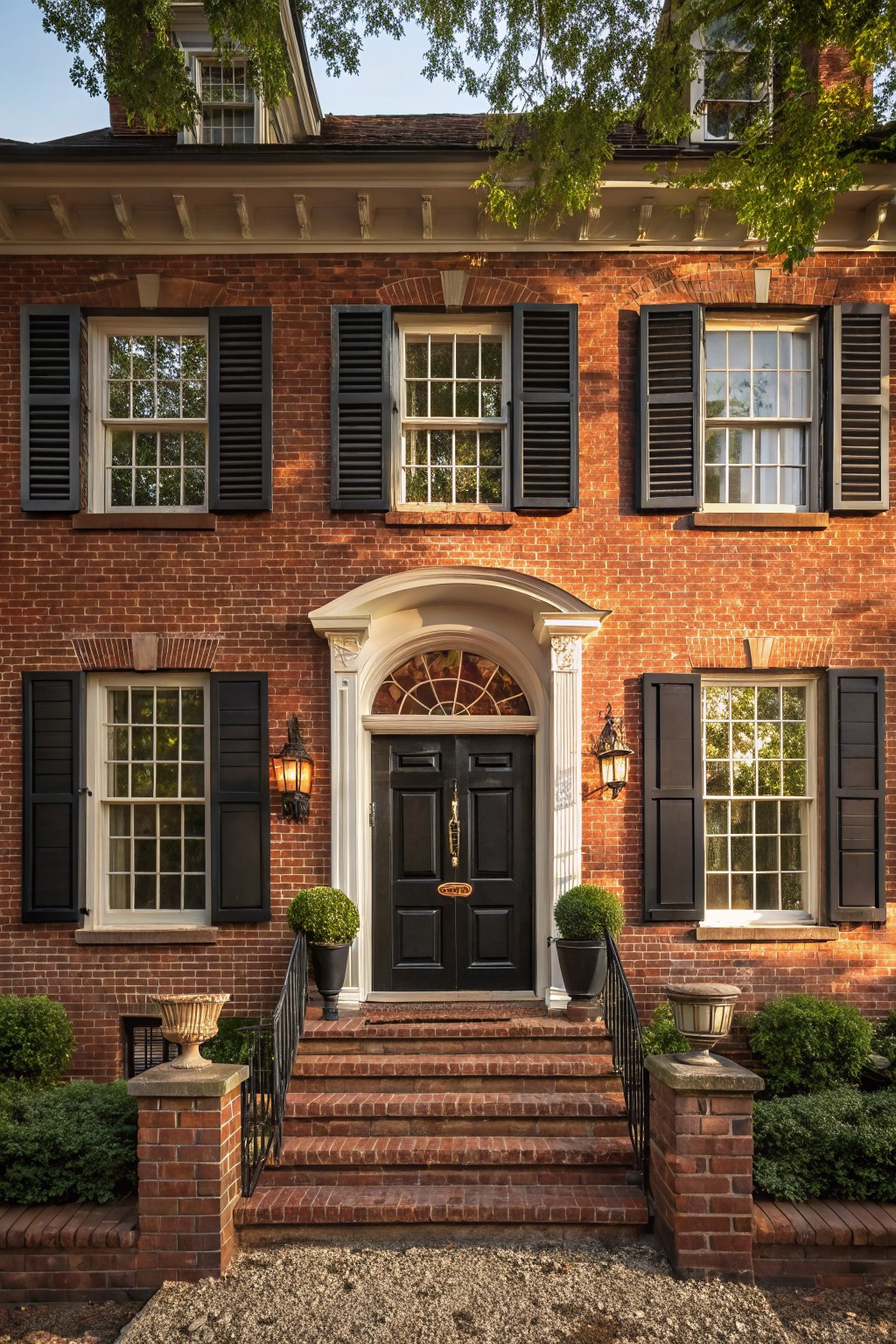 A red brick house exterior with black louvered shutters on white multi-pane windows, a centered black front door under a fanlight transom, flanked by lanterns, brick steps with iron railing, and boxwood topiaries in pots.
