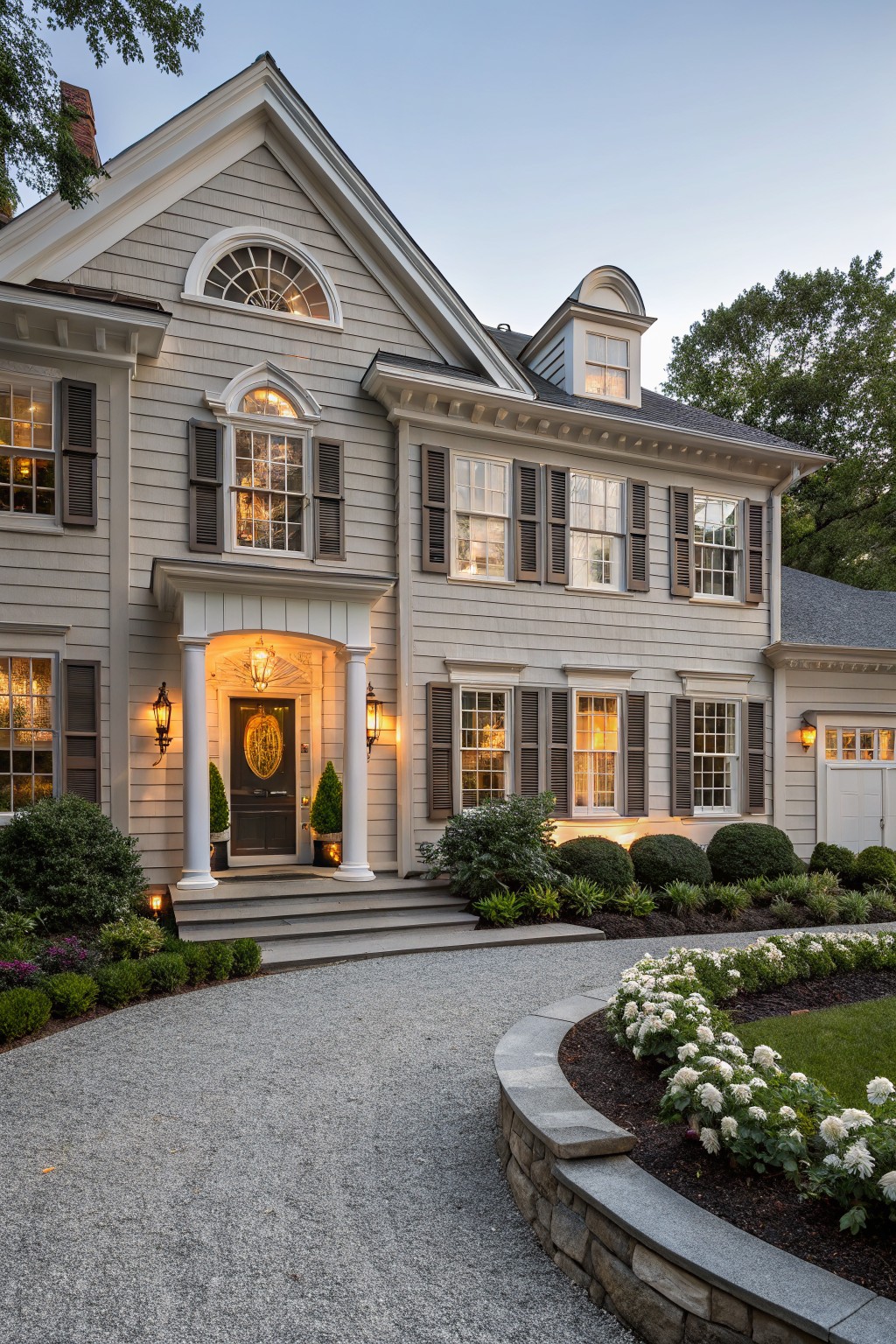 A two-story house with light gray shingle siding, dark brown shutters on multipane windows, a columned front porch with black door and lanterns, boxwood shrubs, and a curved gravel driveway edged in white flowers at dusk.