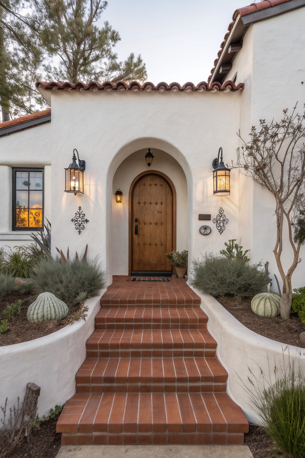 White stucco house facade with arched wooden front door, iron-studded details, wall lanterns, red brick steps leading up, and low desert plants in raised beds.