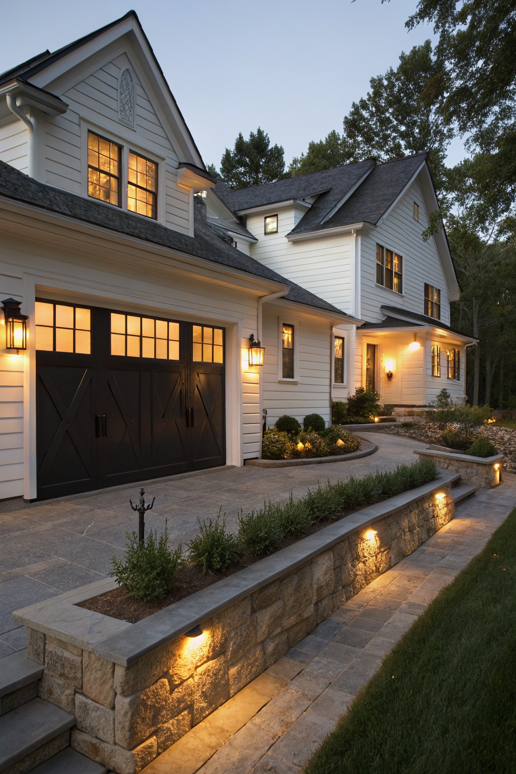 White clapboard house exterior at dusk featuring dark paneled garage doors, lit lanterns, stone retaining wall pathway with plants, and surrounding trees.