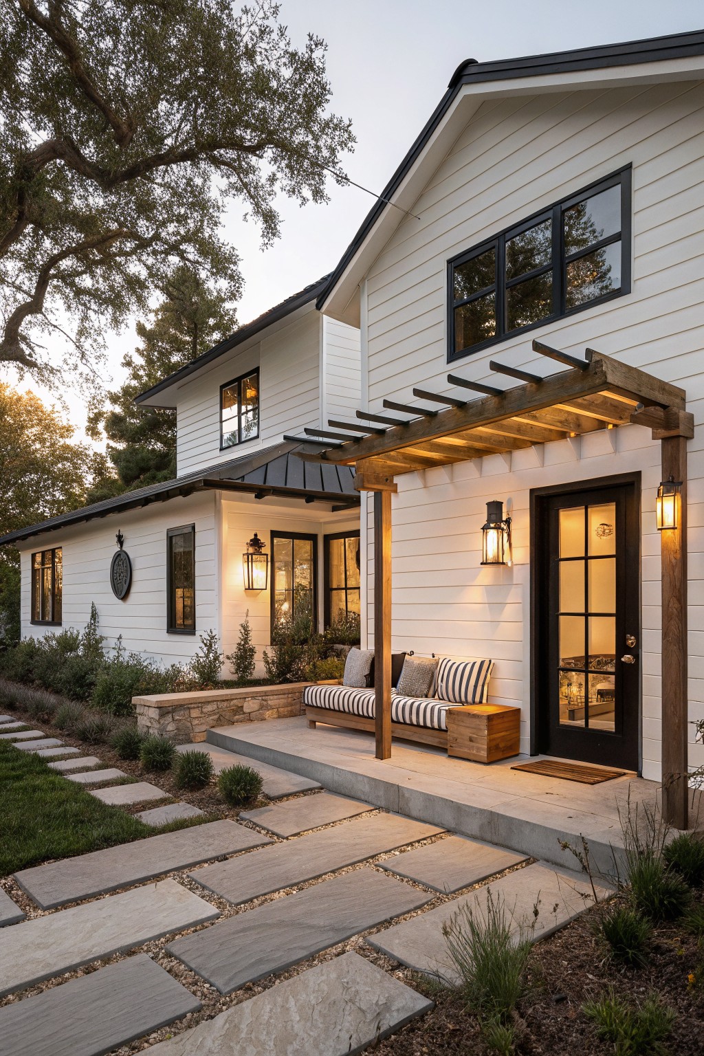 White ranch-style house exterior featuring a wooden pergola over the entry porch with black door, cushioned bench, lantern lights, stone pathway, and low garden plantings at dusk.