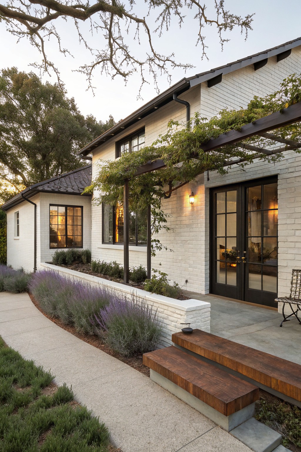 White brick ranch house exterior with a vine-covered black metal pergola over black-framed glass entry doors, wooden steps on a concrete landing, lavender plantings along a curved concrete path, and trees in the background.