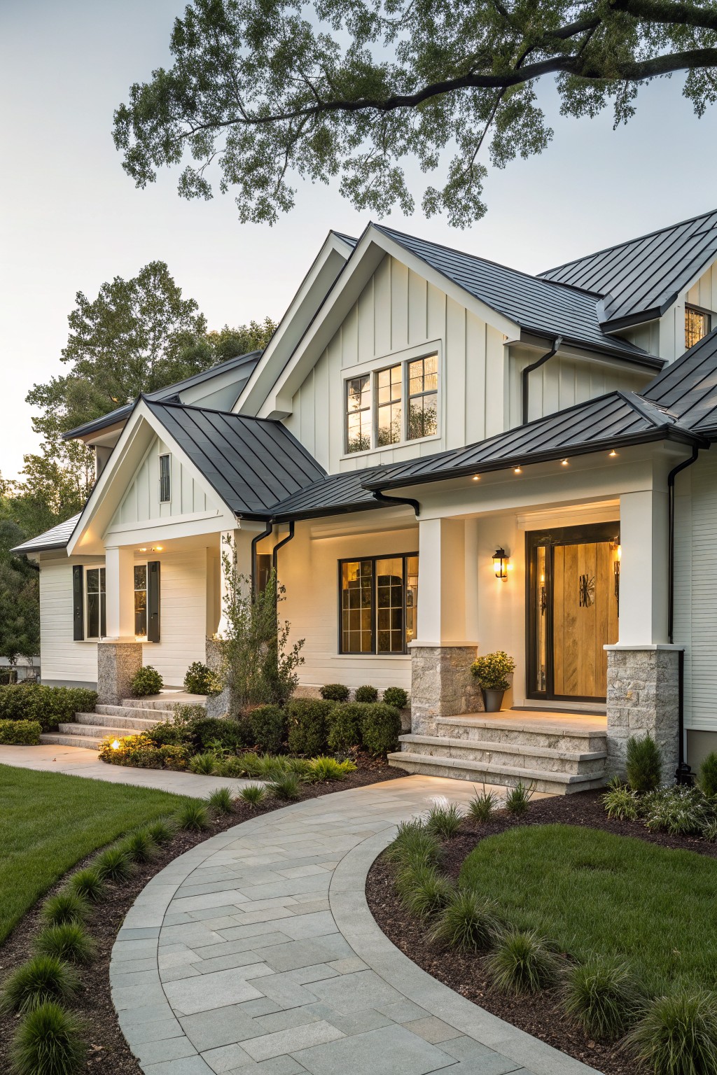 A two-story white board-and-batten house with dark metal gable roofs, stone-columned covered entry porch with wood door, curved stone pathway, shrubs, and lawn at dusk.