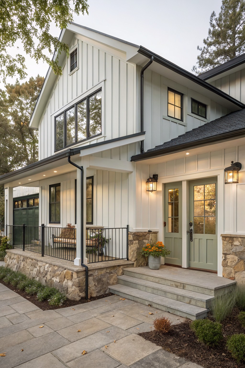 Front exterior of a white board-and-batten house with gabled roof, black trim, covered porch, green double doors, stone foundation, metal railing, and gravel path with plants.