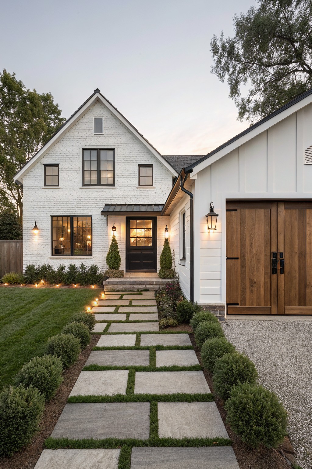 White brick ranch house exterior with gabled roof, black front door flanked by lanterns, attached garage with board-and-batten siding and wood doors, stone pathway with grass joints lined by boxwood shrubs, and lawn in front.