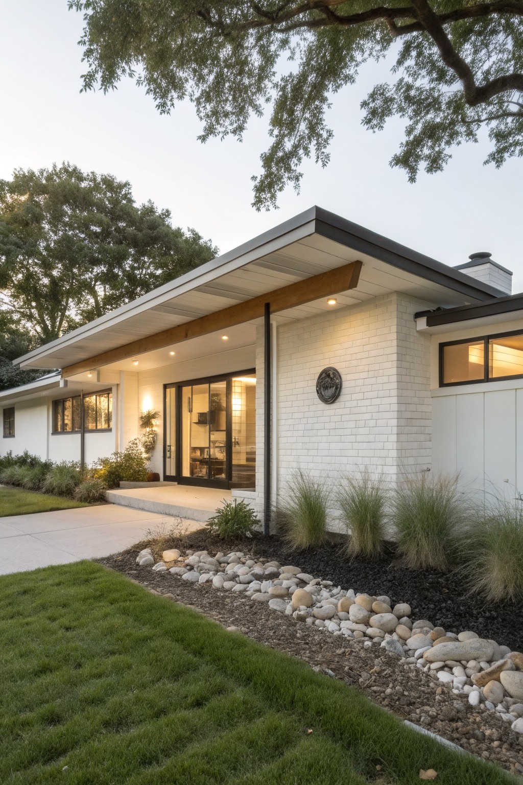 White brick ranch house exterior featuring an overhanging roof with exposed beams over a sliding glass entry door, flanked by black-framed windows, warm entry lighting, and low landscaping with ornamental grasses and river rocks.