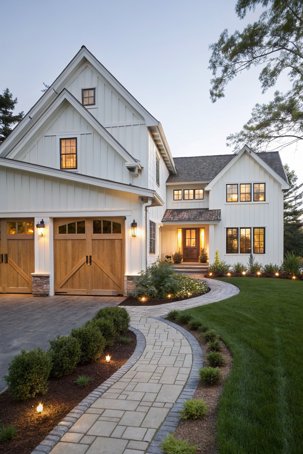 White board-and-batten ranch house exterior at dusk featuring a gabled two-car garage with wooden doors, lit front entry, curved paver pathway, and low shrubs along the driveway.