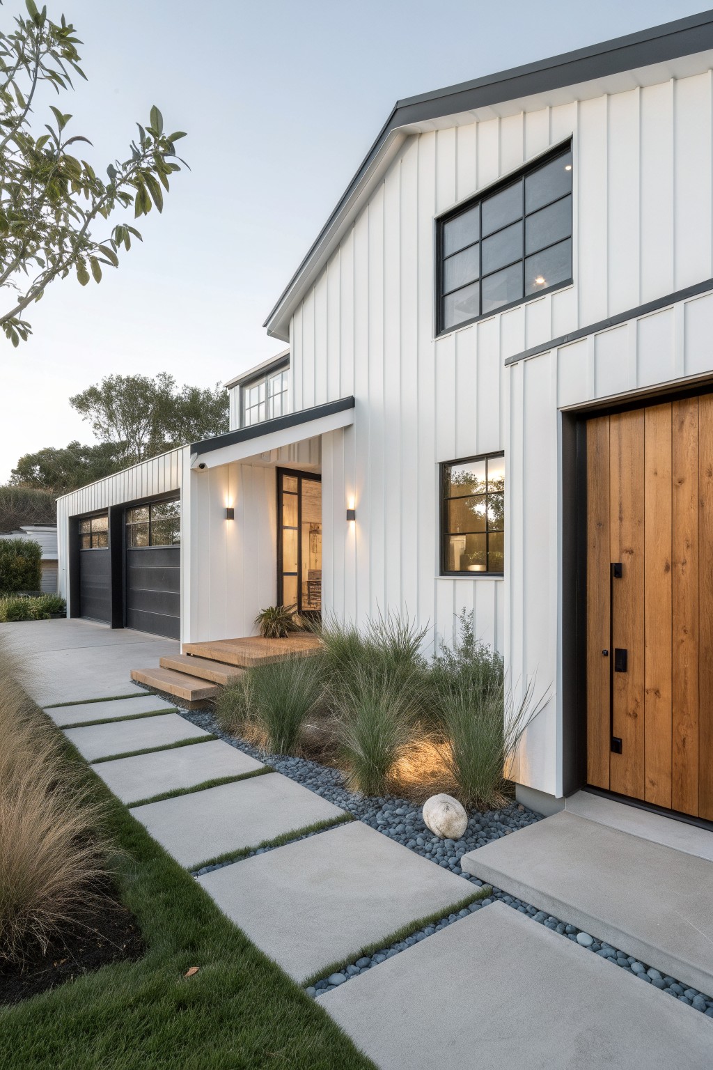 White board-and-batten ranch house exterior featuring a wooden front door, black-framed windows, dark garage doors, concrete paver pathway with gravel strips, and ornamental grasses along the front yard.