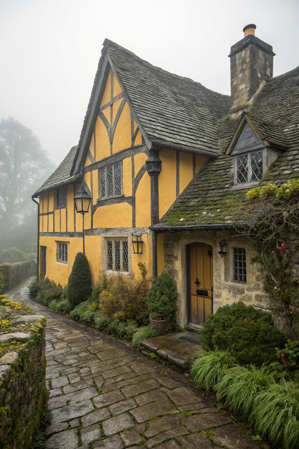 Yellow half-timbered house with gray slate roof, dark timber framing, stone pathway, lanterns, shrubs, and climbing plants in misty weather.