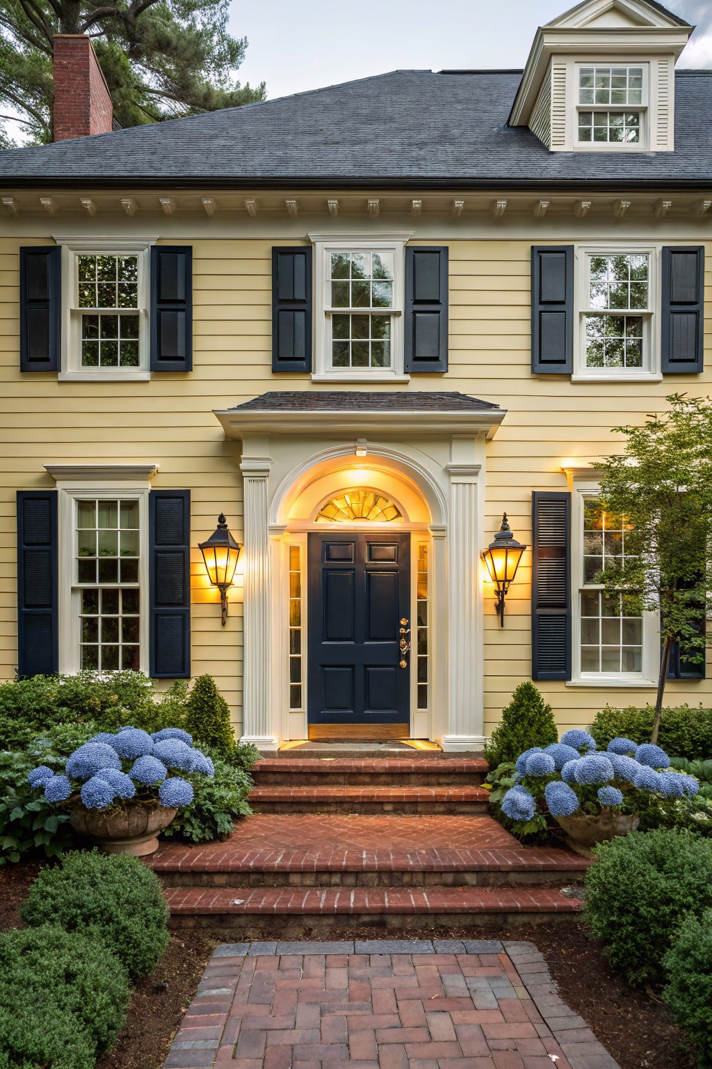 A two-story yellow clapboard house with black shutters, white trim, dark blue front door, lanterns flanking the entry, brick steps, and potted blue hydrangeas.