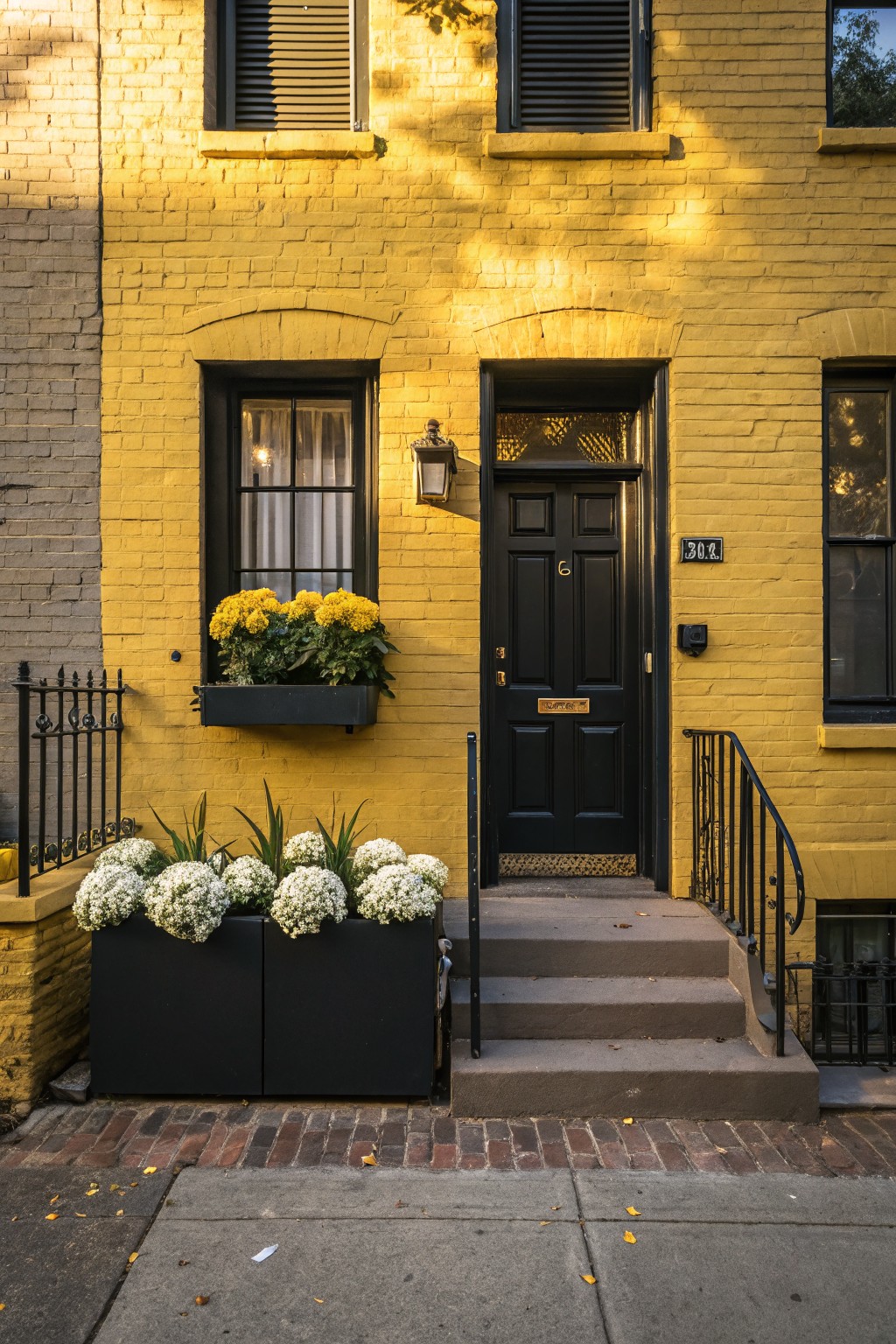 Bright yellow brick townhouse facade with black front door numbered 304A, black window frames, flower boxes holding yellow chrysanthemums and white hydrangeas, black metal railing, and concrete steps.