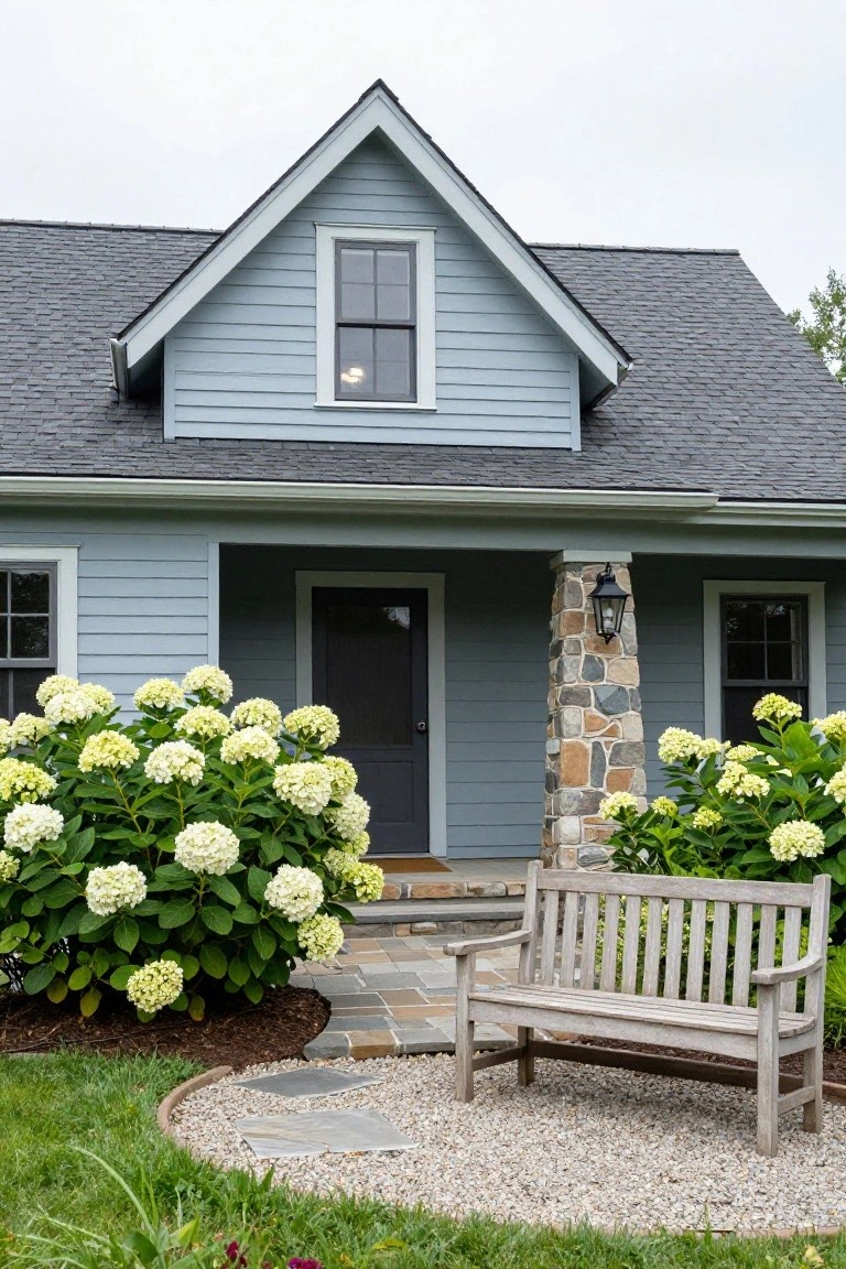 Quaint blue house with hydrangeas, stone pillar, and a wooden bench in the garden.