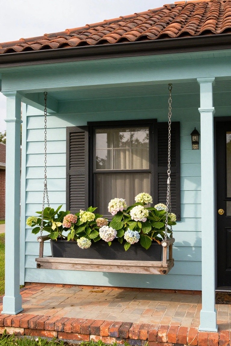 Light blue-green sided house with terracotta tile roof, featuring a hanging wooden porch swing planter filled with white hydrangeas next to the black front door, black shutters on the window, and white porch columns.