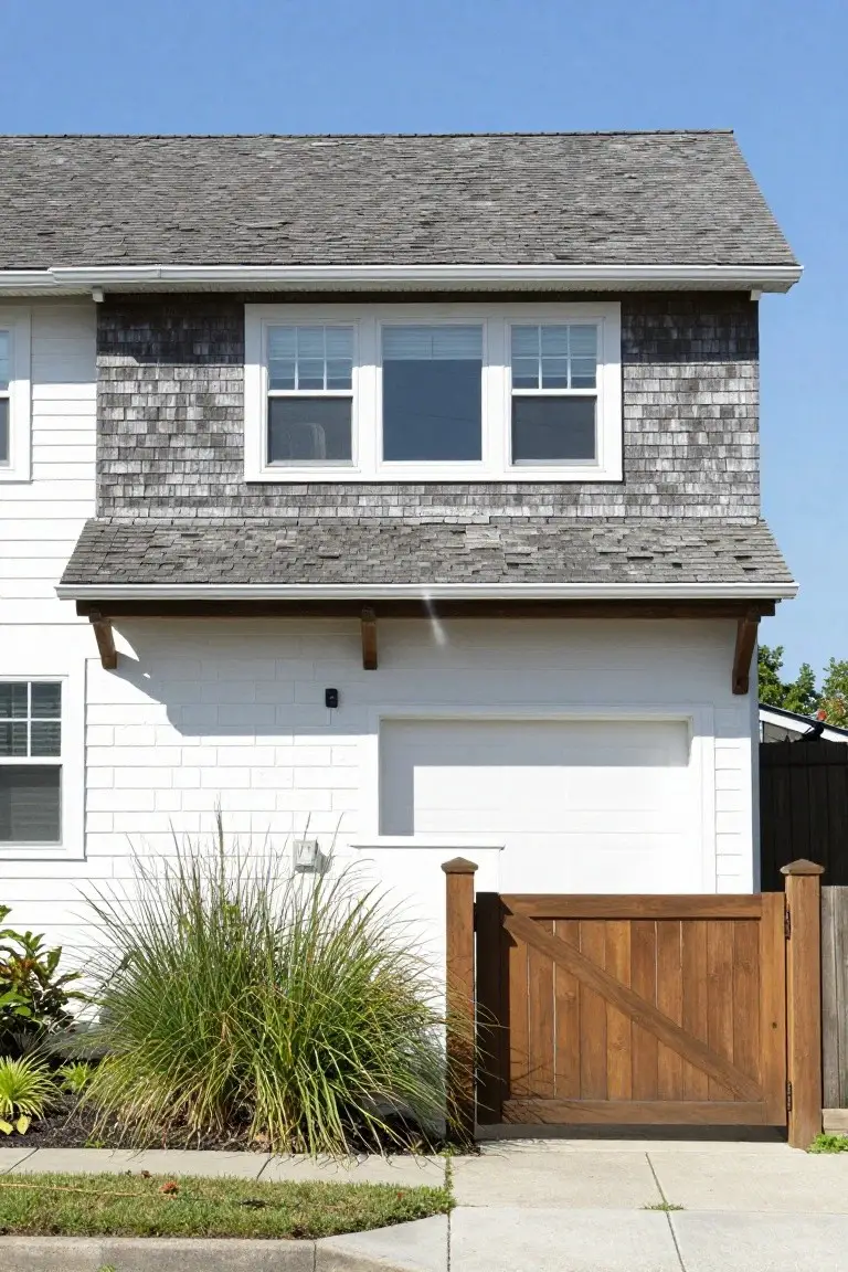 Side view of a two-story white clapboard house with gray cedar shake siding on the upper section and garage overhang, featuring large windows, a white garage door, wooden pedestrian gate, and ornamental grasses in front.