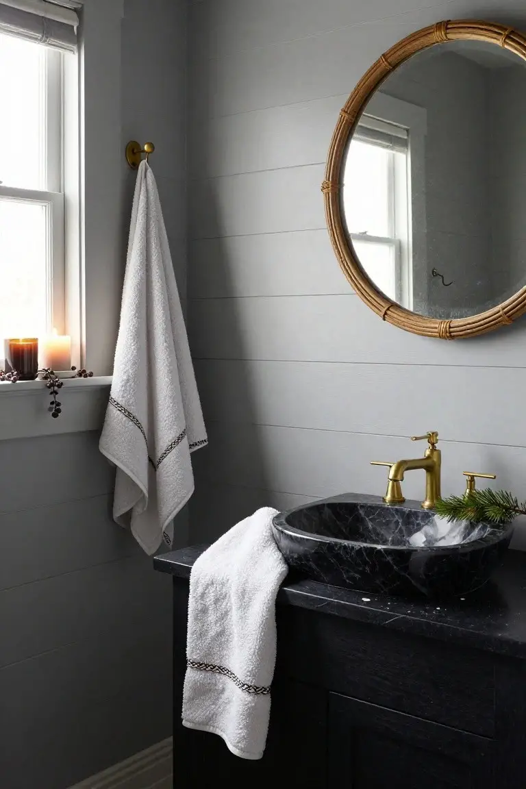 Gray shiplap bathroom with black marble vessel sink on dark wood vanity, gold faucet and handles, rattan mirror, white towels on hook and counter, lit candles and berries on windowsill, pine sprig with white flecks on sink.
