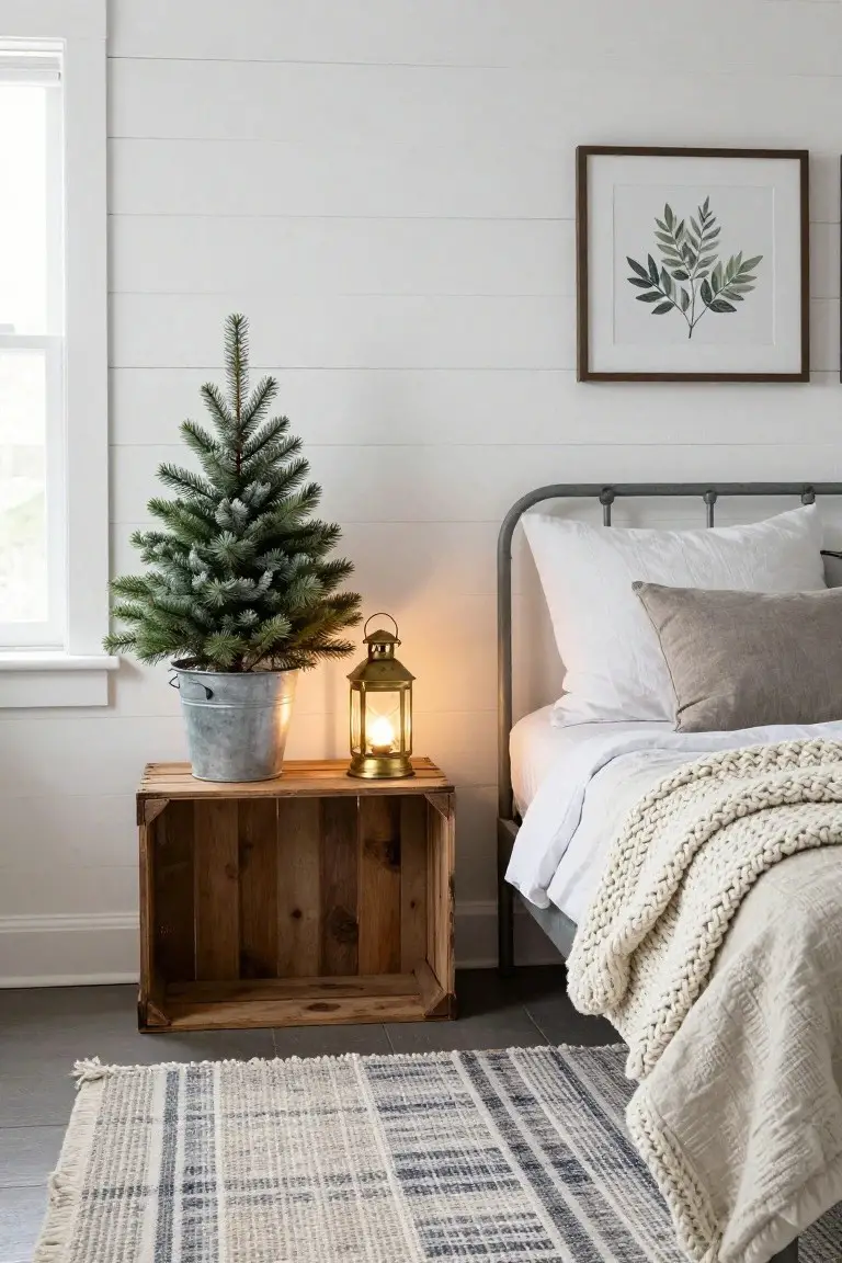 Small artificial flocked Christmas tree in a galvanized metal bucket next to a wooden crate nightstand with a brass lantern on top, beside a gray metal bed frame with white pillows and knitted throw in a white shiplap-walled bedroom.