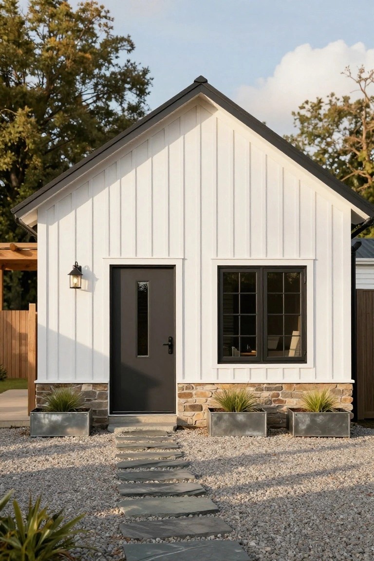 Small white board-and-batten house exterior with black front door and window frames, lantern light beside door, stone foundation with metal planters holding grasses, gravel ground and stone path leading to entry.