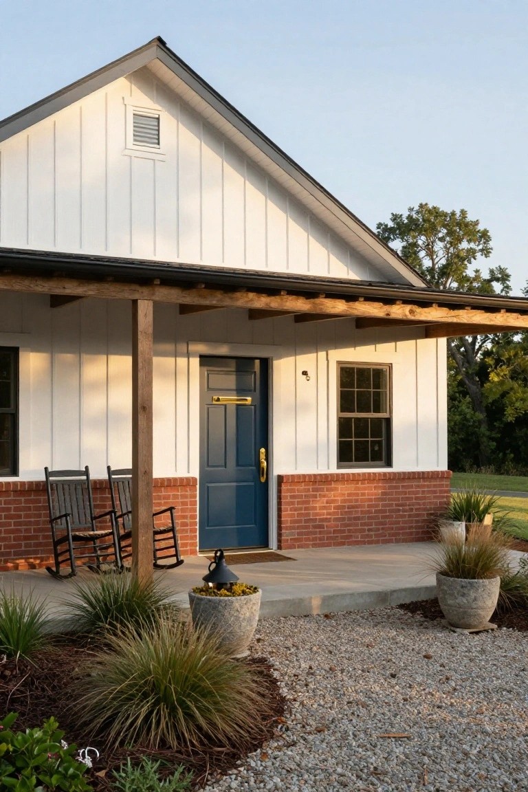White board-and-batten sided house with gabled roof, navy blue front door with brass knocker, red brick accent wall, wooden porch posts, two black rocking chairs, stone lantern, potted grasses, and gravel path.