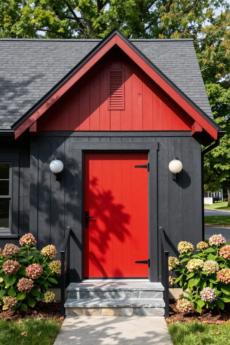 Dark gray shiplap-sided gable building with red front door, black frame, white globe wall lights, hydrangea bushes on either side of gray stone steps, and concrete walkway.