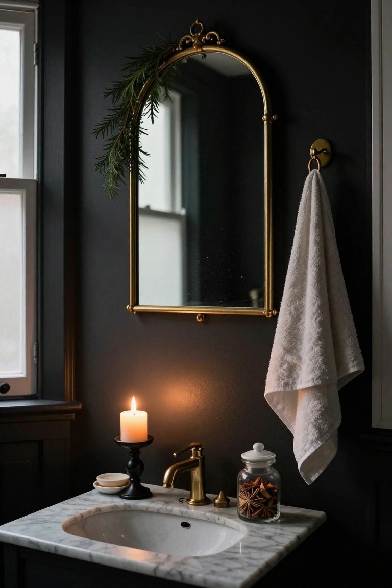 Dark-walled bathroom vanity area featuring a gold arched mirror with evergreen garland draped over the top, a white towel on a gold hook, lit white candle on a black candlestick, glass jar of star anise on marble countertop, and brass sink faucet.