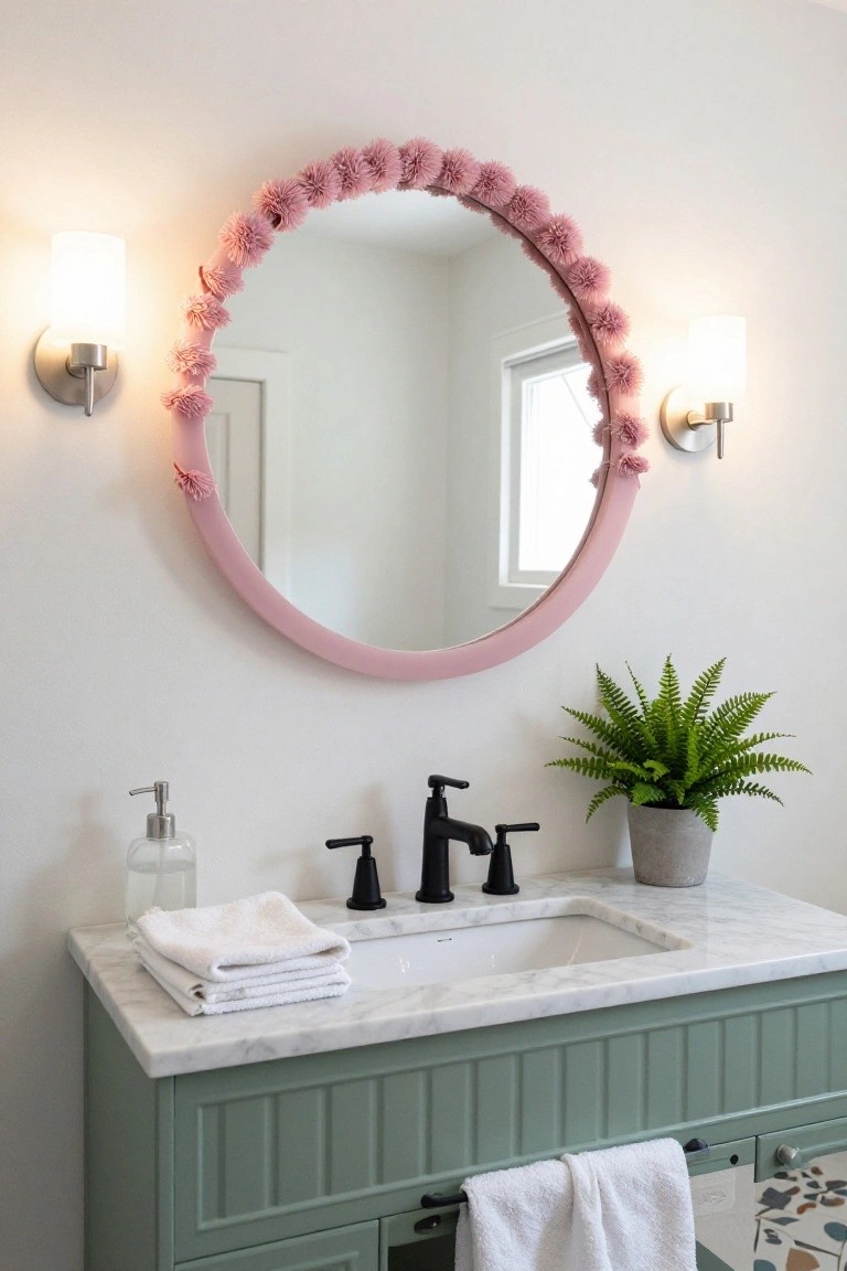 Round bathroom mirror framed with pink pom-pom fringe above a white marble sink on a green shaker-style vanity, flanked by two white globe sconces, with a potted fern plant, soap dispenser, and folded white towels.
