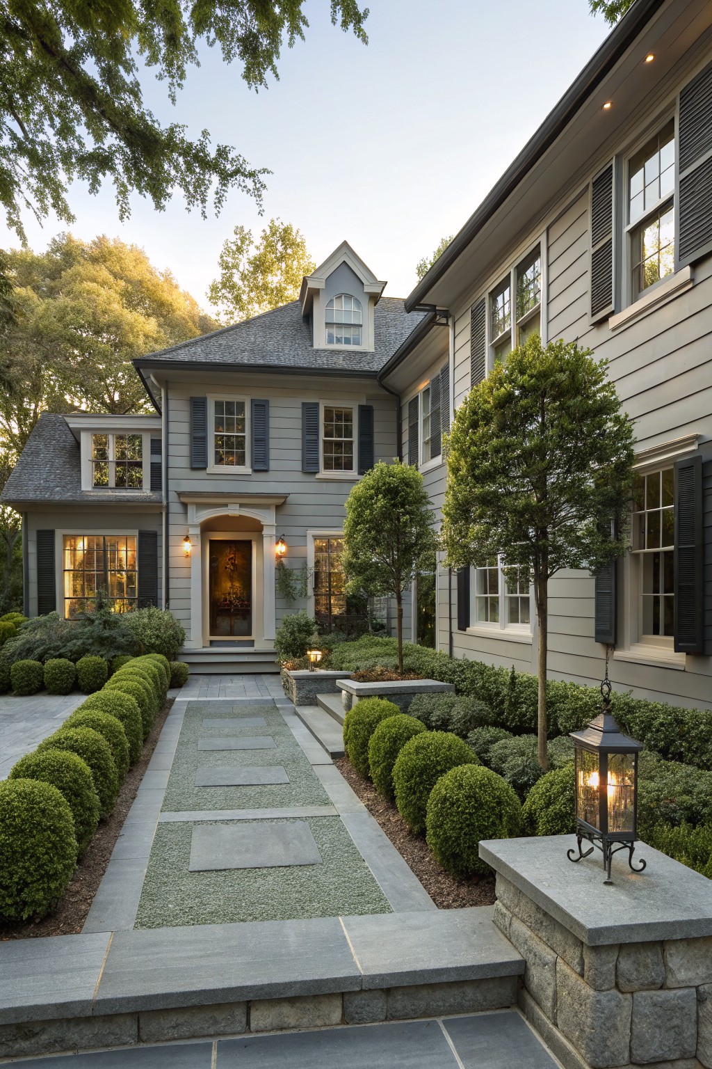Gray shingled house with light gray siding and black shutters, surrounded by trees, featuring a straight stone pathway lined with spherical boxwood shrubs leading to the front entrance, with lanterns on stone pillars.