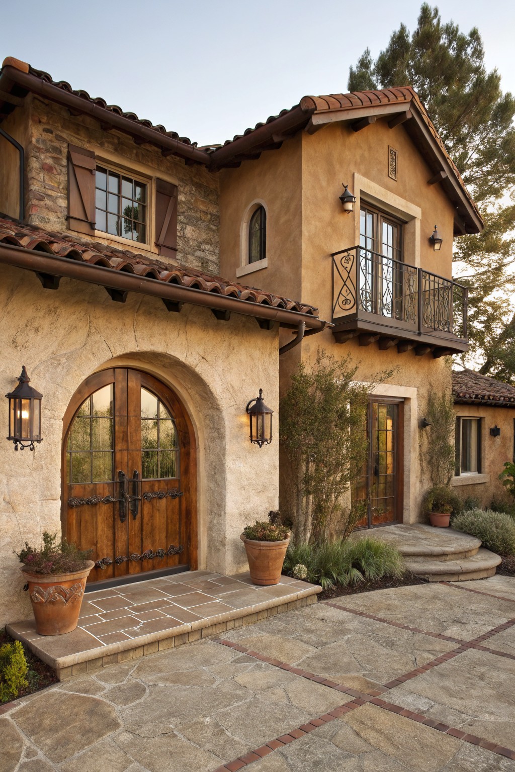 Beige stucco house exterior with red terracotta tile roof, large arched wooden double front door with iron hardware, wrought-iron balcony above, wall lanterns, potted plants, and stone entry steps.