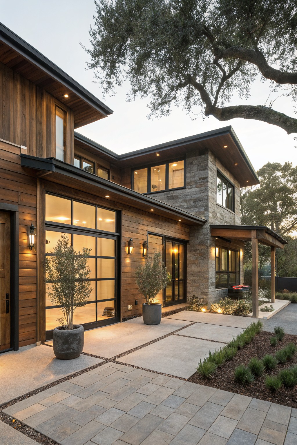 Two-story modern rustic house exterior with dark vertical wood siding, gray stone on a corner tower, large glass garage door, potted olive trees by the entry, stone paver pathway, and mature oak tree branches overhead at dusk.