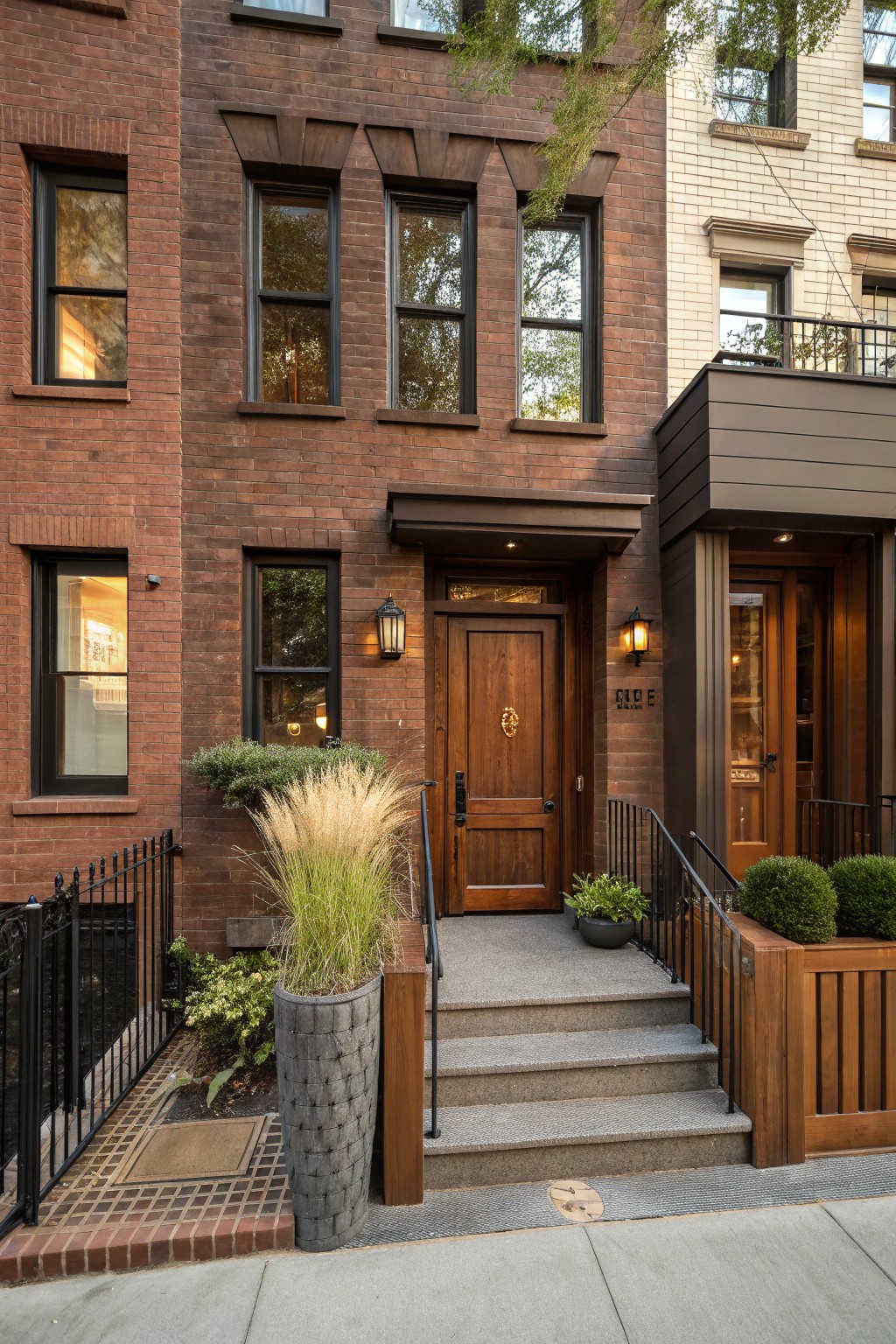 Brown brick row house exterior with wooden front door, brass knocker, wood entry frame, lanterns, stone steps, iron railing, potted grasses and shrubs.