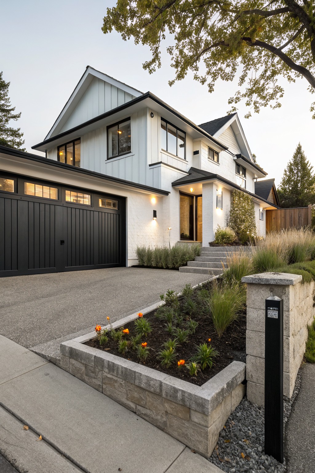 A two-story white shiplap house exterior with black paneled double garage doors, concrete driveway, stone retaining wall planted with orange flowers and grasses, steps to a lit entry, and trees in the background.