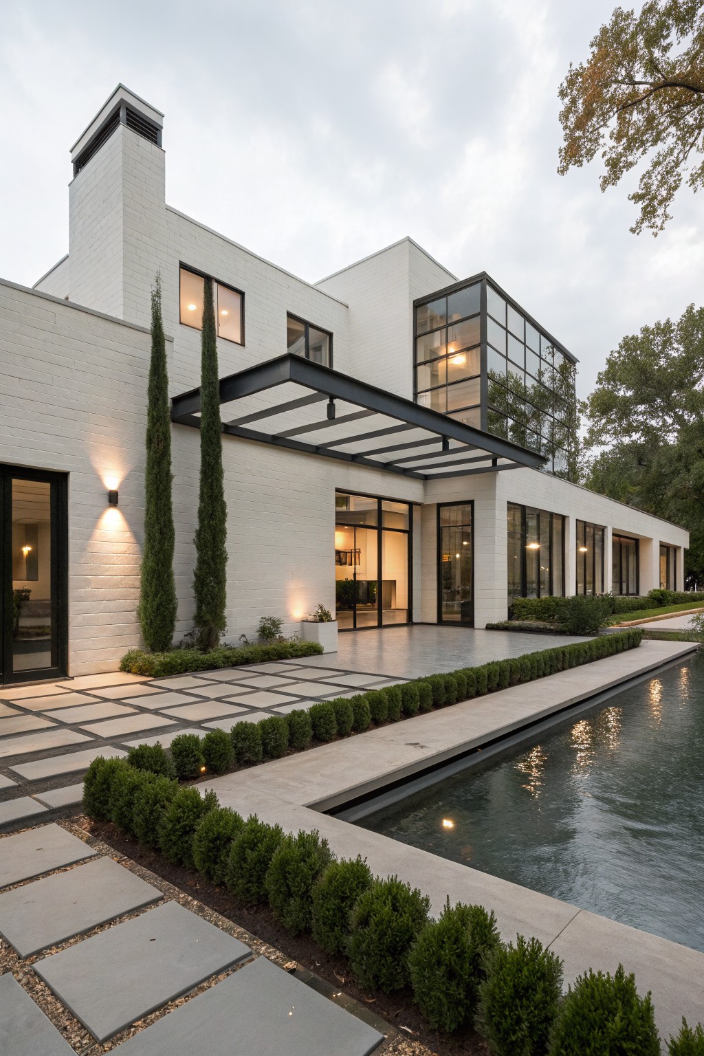 Modern white brick house exterior with a black steel cantilevered canopy over glass entry doors, tall cypress trees flanking the entry, large glass windows, boxwood hedges, and a rectangular reflecting pool in the foreground.