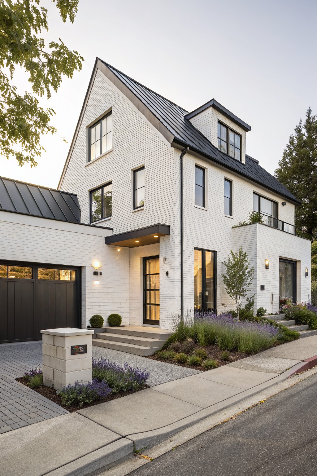 A two-story white brick house with black metal roof, attached black garage, entry porch, and landscaping including lavender plants along the driveway.