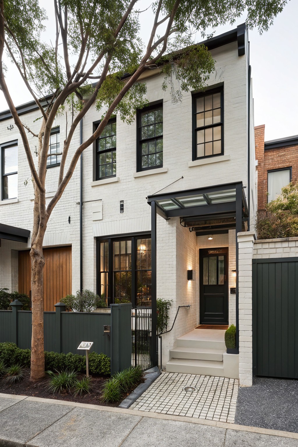 A modern two-story white brick townhouse exterior with black metal window frames, wooden garage door, dark green front door under a glass canopy, green fence, tree, and low plants along the sidewalk.