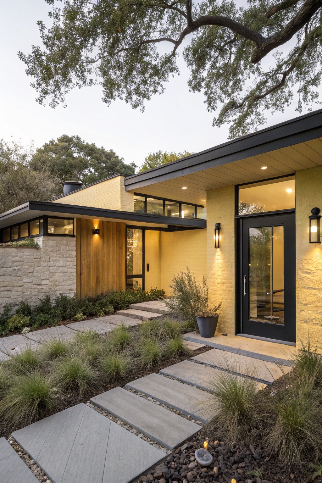 Modern yellow stucco house exterior with black front door, cantilevered roof overhang, wood siding panel, stone accent wall, concrete steps, and gravel path edged by grasses.